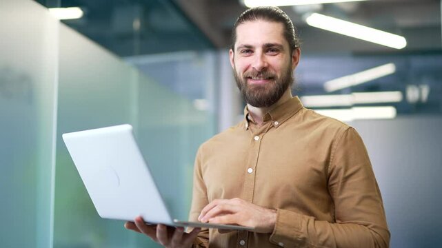 Portrait of a smiling admin IT engineer works on a laptop computer standing in business office of data center. Confident developer technician businessman posing and looking at the camera. Close up