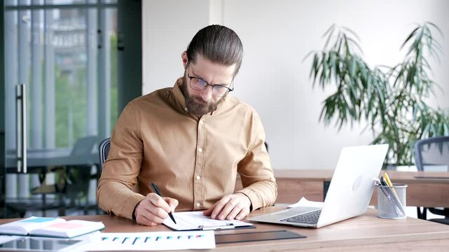 Busy businessman fills out documents with a pen using laptop computer sitting at a work desk at workplace in business office. Male financier doing paperwork, financial report, engaged in accounting