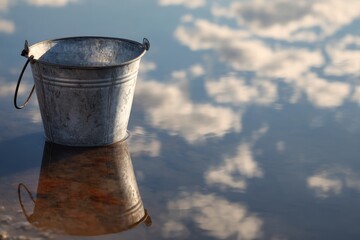 Weathered metal pail standing in shallow water, the sky with fluffy clouds reflected in calm liquid surface
