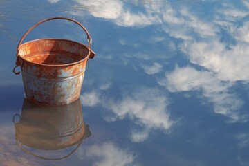Naklejka premium Dilapidated metal bucket sits near water, reflecting cloudy sky, showcasing aging and environmental themes.
