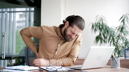 Tired businessman suffering from back pain while working on laptop while sitting at desk at workplace in business office. Upset worker massages painful lower back muscles. He is exhausted, overworked