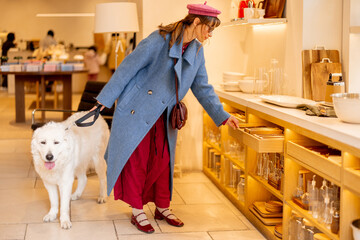 A woman with a pink beret and blue coat walks her dog in a modern boutique, surrounded by stylish shelves. Ideal for commercial use related to fashion, pets, and upscale retail