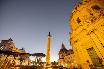 Evening view of Romes iconic Trajans Column and Altare della Patria, illuminated against the twilight sky, surrounded by ruins, churches and cobblestone streets