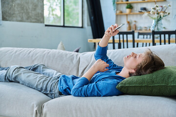 Serious teenager guy looking at smartphone screen lying on the couch at home