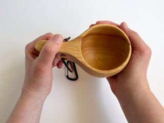 Hands holding a traditional wooden Kuksa cup. Close-up of a handcrafted Nordic mug. Natural wood drinking vessel with a handle and lanyard. Camping and outdoor equipment concept.