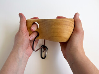 Hands holding a traditional wooden Kuksa cup. Close-up of a handcrafted Nordic mug. Natural wood drinking vessel with a handle and lanyard. Camping and outdoor equipment concept.