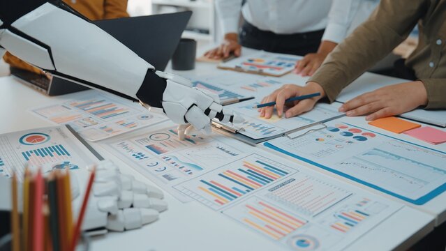A robotic arm aids a business team in analyzing data and developing strategies. The scene depicts modern collaboration with charts, graphs, and documents spread across a table.Noogenesis