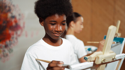 African boy painting canvas with watercolor while happy caucasian girl draw cool tone picture at colorful stained wall. Multicultural highschool student attend creative activity together. Edification.
