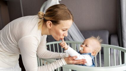 Young mother gently Kissing baby daughter in crib in bright nursery. Tender parenting moment, bonding, care and love at home. Real indoor lifestyle scene with soft light and warmth