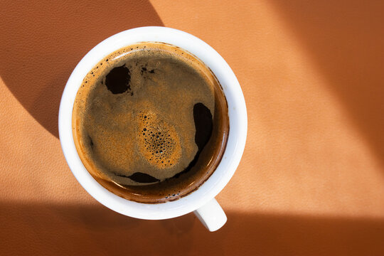 Overhead view of hot black espresso coffee with rich crema in a white cup on a brown table with sharp morning sun shadow for cafe shop banners, warm drink posters, and modern wallpaper.
