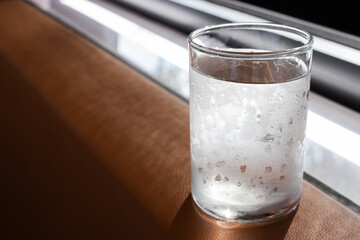 A cold glass of water with condensation sits on a brown surface by the window, catching natural light. The droplets suggest freshness and coolness, ideal for hydration or lifestyle themes.