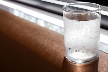 A cold glass of water with condensation sits on a brown surface by the window, catching natural light. The droplets suggest freshness and coolness, ideal for hydration or lifestyle themes.