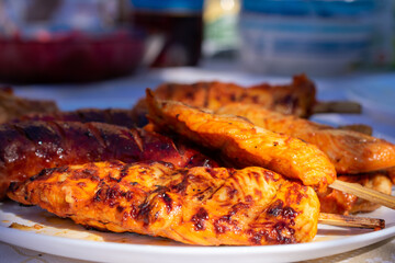 Close-up of grilled meat skewers and sausages on a white plate, served outdoors on a sunlit table, perfect for a summer barbecue.