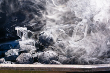 Close-up of charcoal briquettes heating in a grill with white smoke rising. The coals are still mostly black, indicating the early stage of lighting.