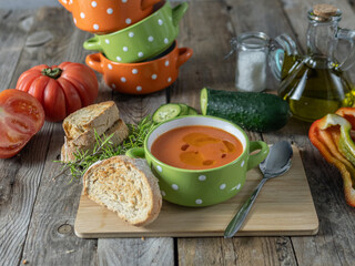 A horizontal photo of a plate of gazpacho in a fun bowl with the ingredients needed to prepare it around it on a wooden table.
