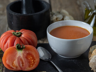 vertical photo of a white bowl of gazpacho on a black slate accompanied by various ingredients and kitchen utensils on a wooden table.
