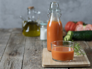A horizontal photo of a glass of gazpacho and a bottle in the background, on a tray isolated on a wooden background, with ingredients in the background. Copy space