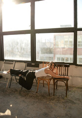 Brunette woman resting across vintage chairs near large window