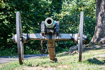 A vintage cannon stands still on a grassy knoll, pointing towards a sprawling battlefield. The sky shines blue, and trees frame the horizon, evoking a sense of history.