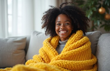 Beautiful black woman smiles sitting wrapped under yellow blanket. Cheerful african american female relaxing at home, enjoying wintertime cozy warmth on couch. Smiling brunette happy person. Winter