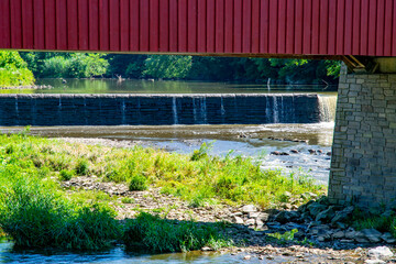 A red covered bridge arches over a serene river where water cascades over a low dam. Lush greenery surrounds the banks, highlighting the beauty of summer in a peaceful rural area.