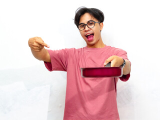 Young Asian man in a pink t-shirt excitedly points to a frypan while holding it. Isolated on a...