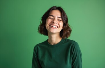 Joyful woman with closed eyes against green background. Attractive female brunette with big smile, white teeth. Portrait of happy girl shows positive emotions. Model wearing dark green clothes in