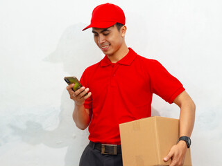 Smiling male courier in red uniform and cap holding a package box and mobile phone, isolated on...