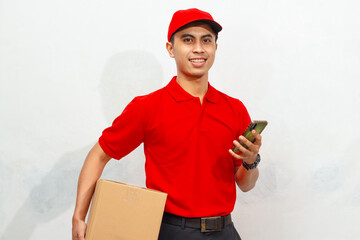 Smiling male courier in red uniform and cap holding a package box and mobile phone, isolated on...