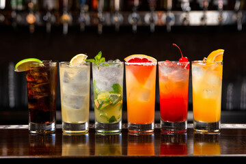 Vibrant lineup of six assorted highball cocktails with fresh fruit garnishes on a dark reflective wooden bar counter under bar lighting
