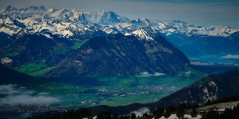 Naklejka premium Majestic snow-capped mountains tower above a serene green valley in Switzerland during a cloudy day