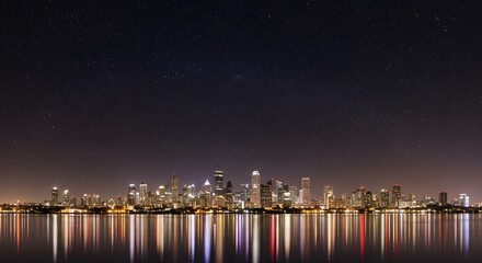 Night city skyline reflecting in water under a starry sky, creating a serene urban landscape with vibrant illuminated architecture.