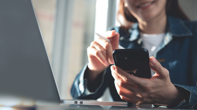 Smiling young asian business woman using mobile phone, working on laptop computer at office. Entrepreneur woman chatting via mobile videochat app, working at home, e-learning