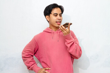 A young Asian man in a pink hoodie happily poses while recording a voice note with his smartphone.