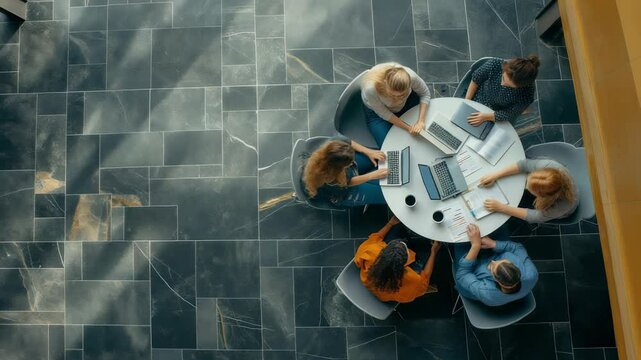 A top-down view shows a group of six diverse individuals collaborating around a white round table with laptops and papers in a workspace featuring grey stone tiled flooring. Focused teamwork.