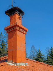 Orange chimney on the roof covered with orange tiles against a background of pines and blue sky on a clear day