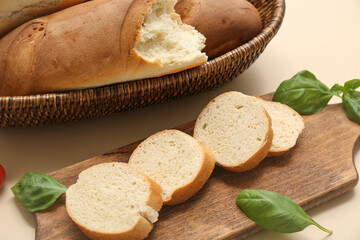 Wooden board and wicker bowl with fresh baguettes and basil on beige background, closeup