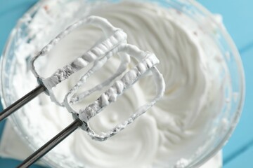 Hand mixer with whisks and bowl of whipped cream on light blue table, above view