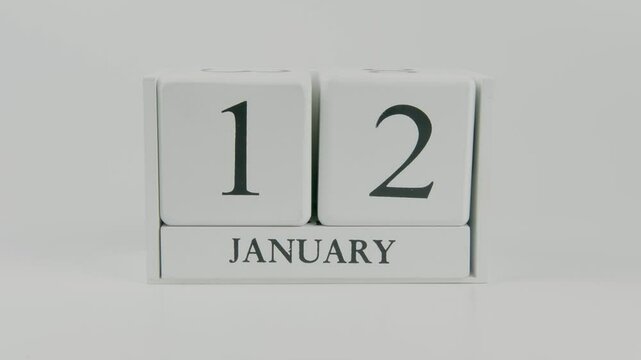 A hand flips a wooden calendar from January 12th to January 13th on a white background, marking the passing of time and the start of a new day