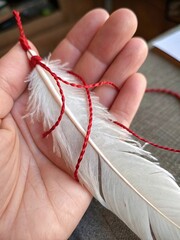 Close up of a hand holding a wrapped white feather with red string in a cozy indoor setting