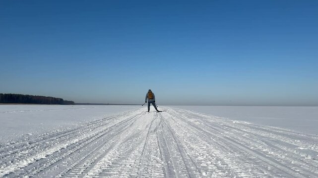 A man is skiing alone at a skating pace along a highway on a frozen reservoir. Caucasian adult skier in a backpack trains in winter. view from the back