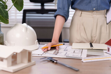 Fototapeta premium Asian Woman Architect Drawing Building Plan With Pencil, Ruler and Hard Hat In Office To Design Construction Project For Building Design With Professional Architecture