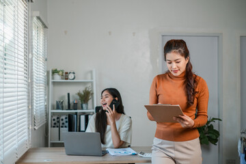 Two women collaborating in a bright office, one using a laptop and phone, the other reviewing documents.