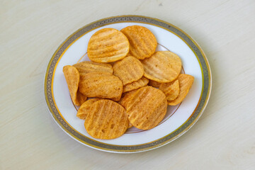 Crispy potato chips on a ceramic plate, resting on a light wooden surface. Delicious Bombay Sweets Alooz Crackers in Charcoal BBQ flavor – wavy perfection!