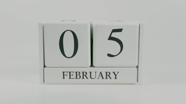 A hand flips a wooden calendar from February 5th to February 6th on a white background, marking the passing of time and the start of a new day
