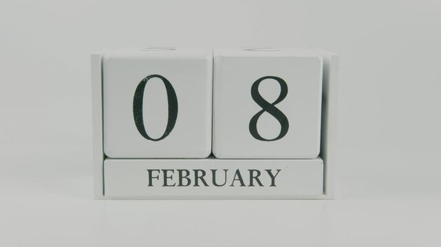 A hand flips a wooden calendar from February 8th to February 9th on a white background, marking the passing of time and the start of a new day
