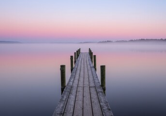 Obraz premium Wooden pier extending into a calm lake at dawn with pink and purple sky reflecting on the water and mist