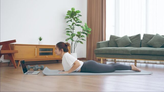 Young Asian woman stretching yoga workout on exercise mat while online training class with computer laptop in living room