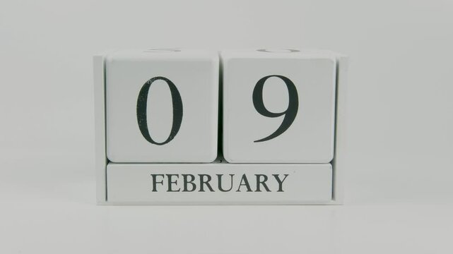 A hand flips a wooden calendar from February 9th to February 10th on a white background, marking the passing of time and the start of a new day