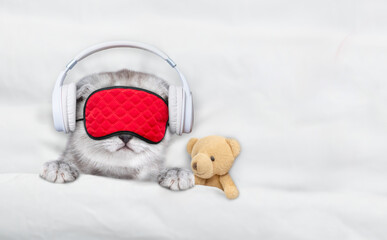 Cute kitten wearing  headphones and mask for sleep listening music with toy bear on a bed at home....